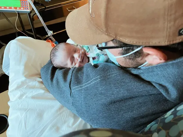 A man in a baseball cap cradling a baby in a hospital bed, creating a heartwarming bond.