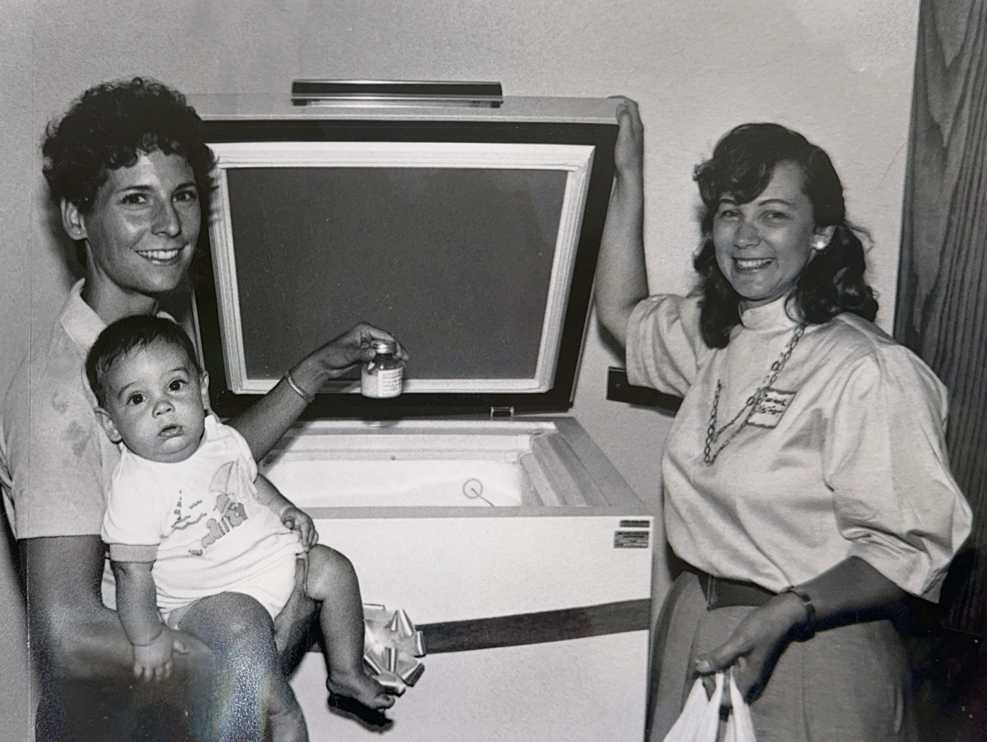 Black and white image of two women standing by a freezer. One is holding a baby and a bottle of donor milk
