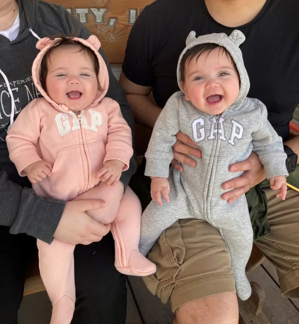 two twin baby girls sitting on their parents' knees wearing onesies with bear ears on them that say 